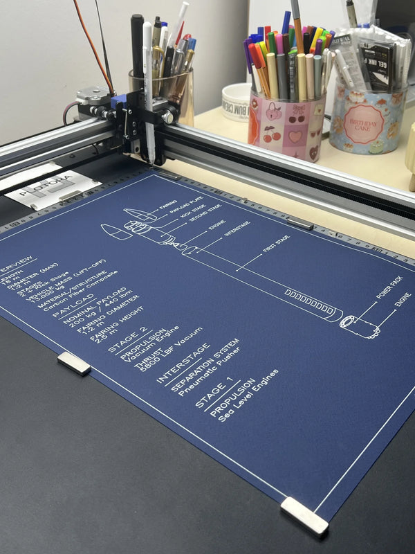 A desktop pen-plotter machine in use, printing a detailed technical schematic onto a large navy blue sheet. The machine's carriage is visible above the sheet, and a desk with pens and other office items sits in the background.