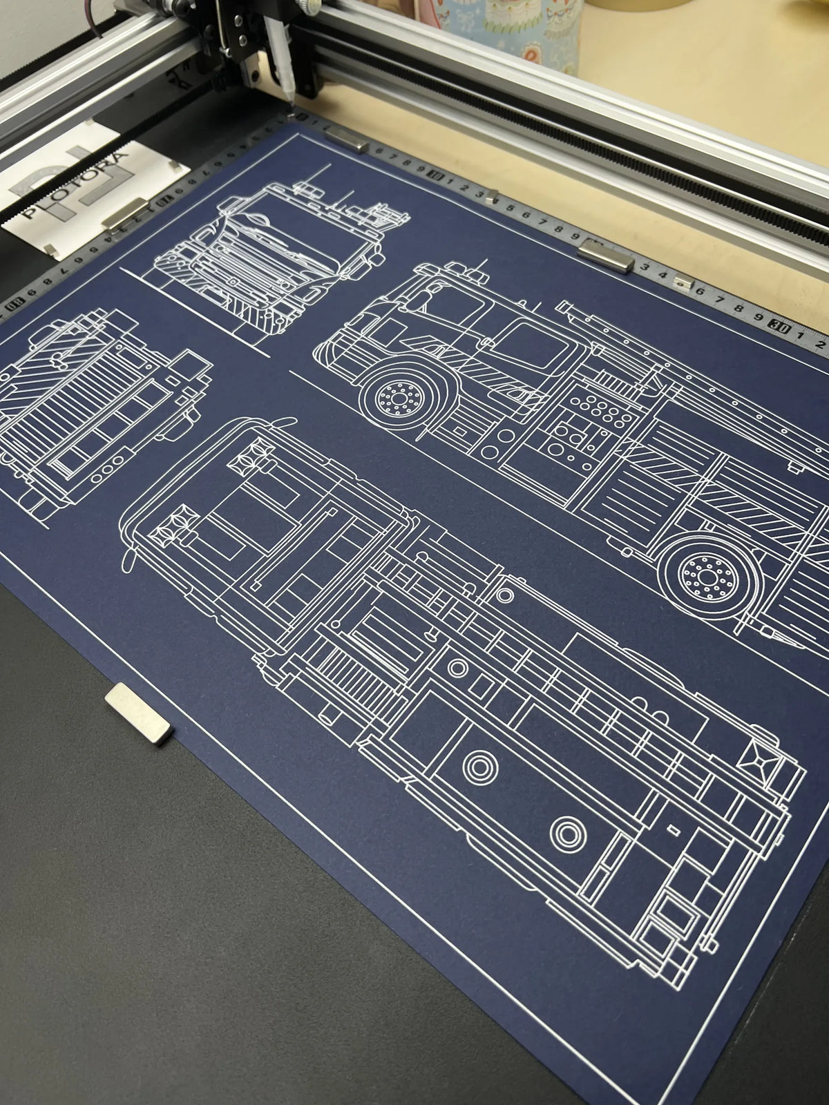 Fire truck blueprint being drawn by a pen plotter in white ink on navy blue paper, showing side, front and top views