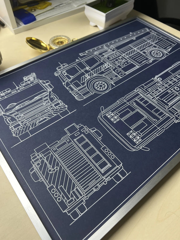 Fire truck blueprint art in a silver metal frame, displayed on a desk with a brass compass