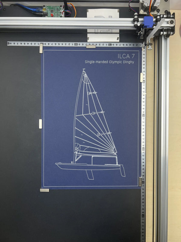 A blueprint-style poster of the ILCA 7 Single-Handed Olympic Dinghy, featuring a white line drawing on a dark blue background, mounted on a black board inside a 3D printer frame.