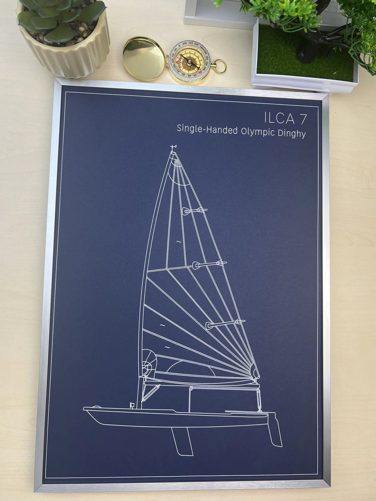 A framed technical blueprint poster of the ILCA 7 Single-Handed Olympic Dinghy, featuring a white line drawing on a dark navy blue background, styled on a light wood surface with decorative plants and a pocket watch.