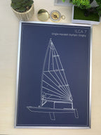 A framed technical blueprint-style print of an Olympic dinghy, labeled 'ILCA 7 Single-Handed Olympic Dinghy,' shown on a light wood surface with small potted plants and a compass nearby.