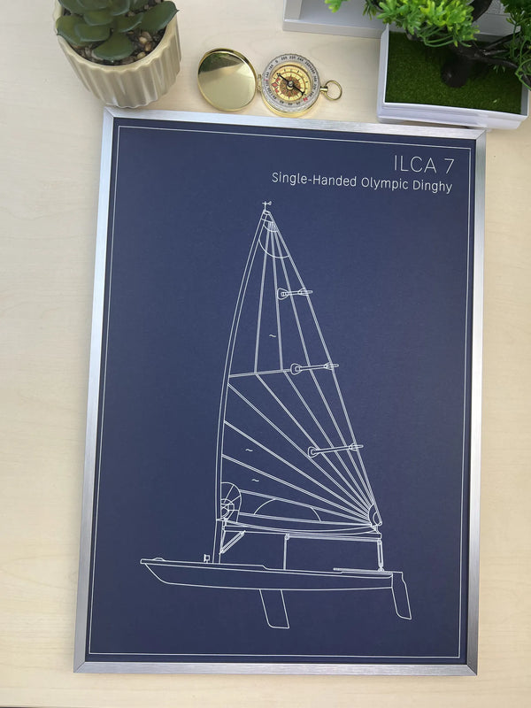 A framed technical blueprint-style print of an Olympic dinghy, labeled 'ILCA 7 Single-Handed Olympic Dinghy,' shown on a light wood surface with small potted plants and a compass nearby.