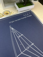 A navy blue printed diagram card titled 'ILCA 7 Single-Handed Olympic Dinghy' with white line drawings of a boat rigging layout, photographed on a tabletop with a small plant and compass nearby.