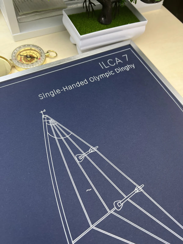 A navy blue printed diagram card titled 'ILCA 7 Single-Handed Olympic Dinghy' with white line drawings of a boat rigging layout, photographed on a tabletop with a small plant and compass nearby.