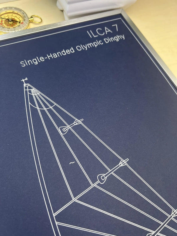 A close-up of a navy blue printed card or booklet cover titled 'ILCA 7 Single-Handed Olympic Dinghy' with white line drawings of a sailboat rigging diagram. A small gold compass is partially visible at the top left on a light surface.