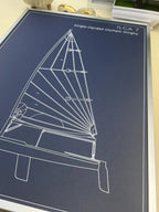 A framed or mounted print featuring a white line drawing of an ILCA 7 single-handed Olympic dinghy on a dark navy background, photographed from above on a light surface.