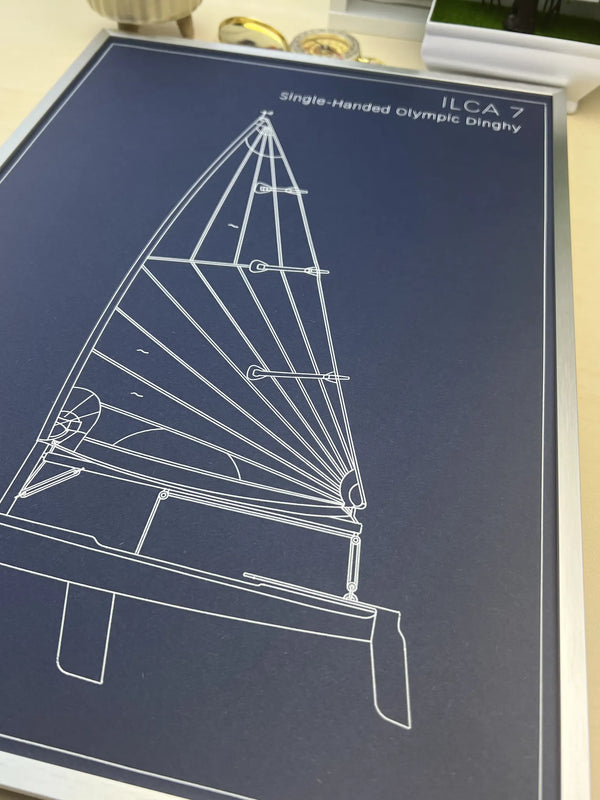 A framed or mounted print featuring a white line drawing of an ILCA 7 single-handed Olympic dinghy on a dark navy background, photographed from above on a light surface.