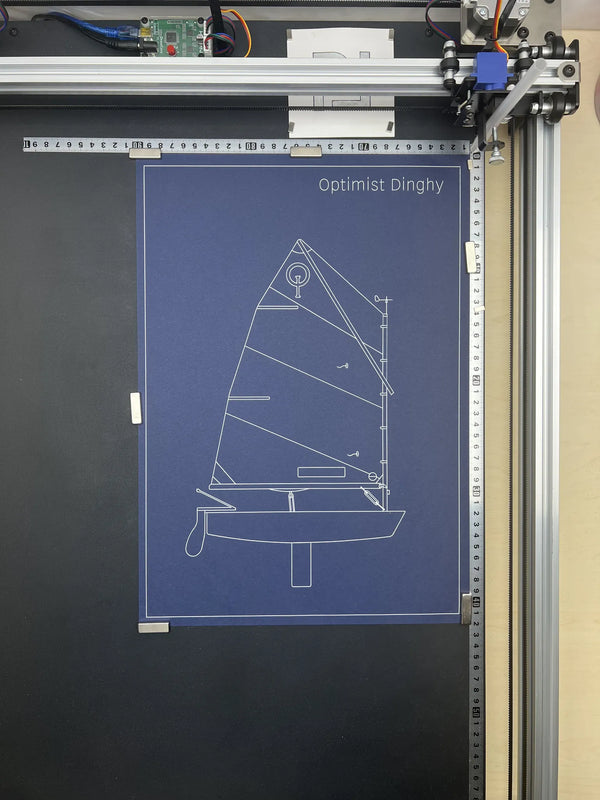 A blue blueprint-style poster featuring the white line drawing and title 'Optimist Dinghy' is mounted on a dark wall, photographed in a workshop setting with a 3D printer frame visible above and to the right.