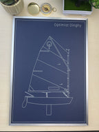 A framed blueprint-style poster of a dinghy sailboat titled 'Optimist Dinghy' in white line art on a dark blue background, displayed on a light wood surface with small decorative objects around it.