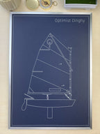 A framed art print featuring a white line drawing of a sailboat on a dark navy blue background, with the text 'Optimist Dinghy' at the top. The print is shown framed and resting on a light beige surface.