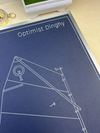 A close-up of a printed poster or art print titled 'Optimist Dinghy' with white line drawings on a dark blue background, placed on a light wood surface.