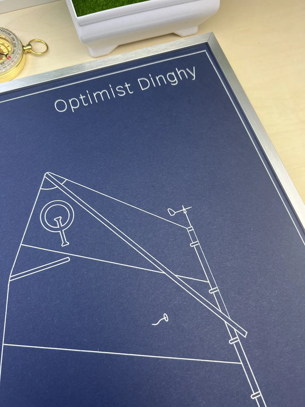 A close-up of a printed poster or art print titled 'Optimist Dinghy' with white line drawings on a dark blue background, placed on a light wood surface.