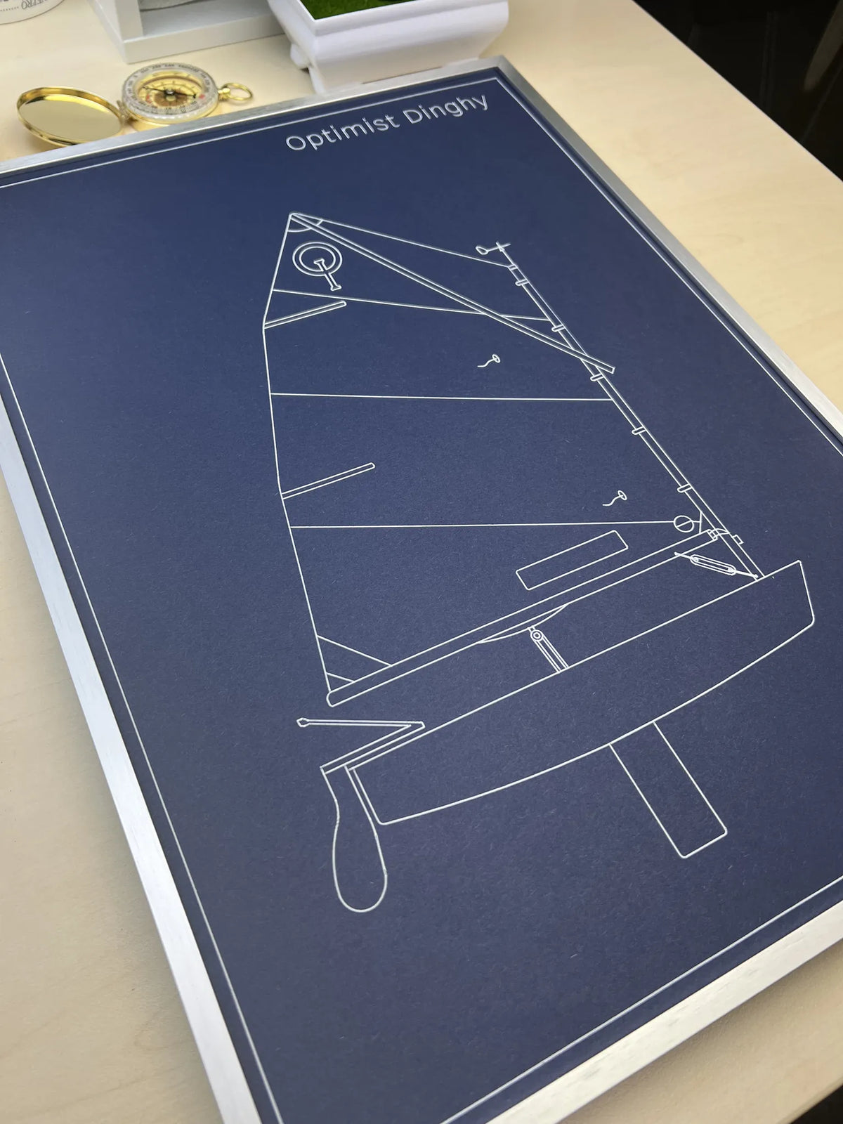 A framed art print featuring a white line drawing of a sailboat on a dark navy blue background, titled 'Optimist Dinghy'. The print is displayed on a light-colored wooden desk with a gold pocket watch nearby.