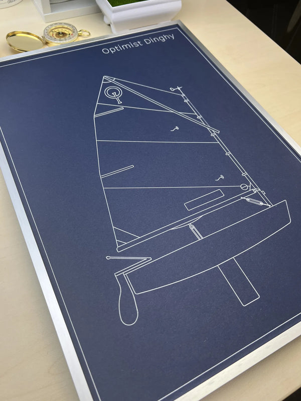 A framed art print featuring a white line drawing of a sailboat on a dark navy blue background, titled 'Optimist Dinghy'. The print is displayed on a light-colored wooden desk with a gold pocket watch nearby.